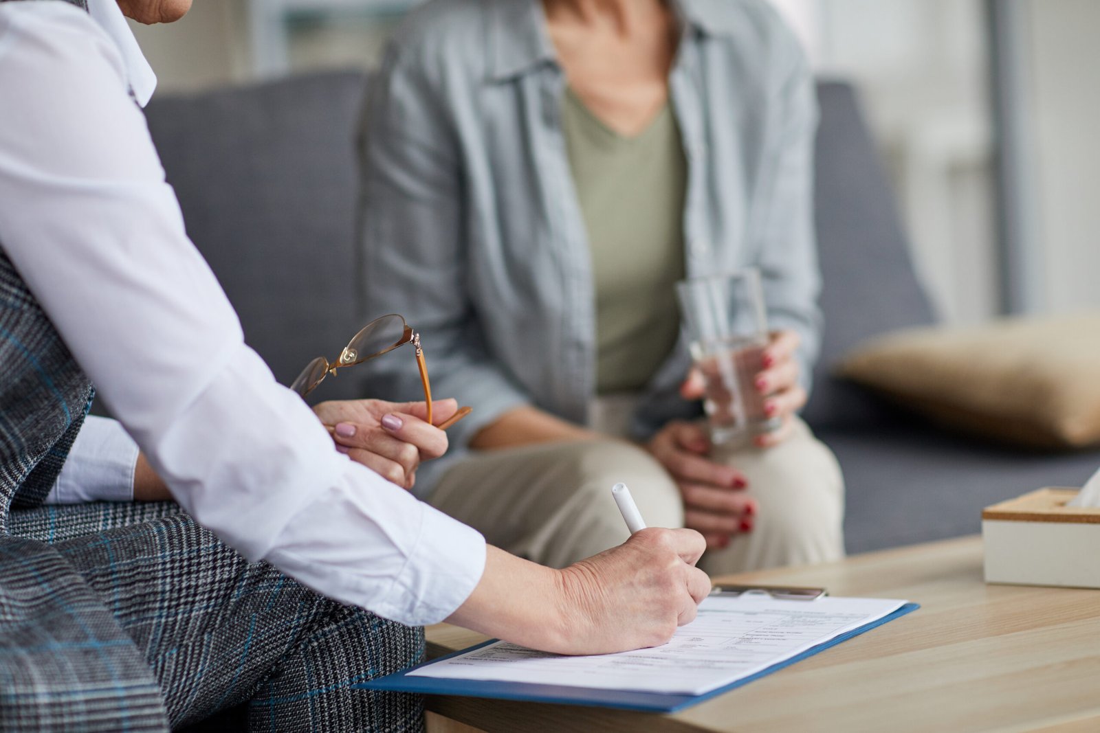 Close up of unrecognizable female psychologist writing on clipboard while talking to mature woman during therapy session, copy space
